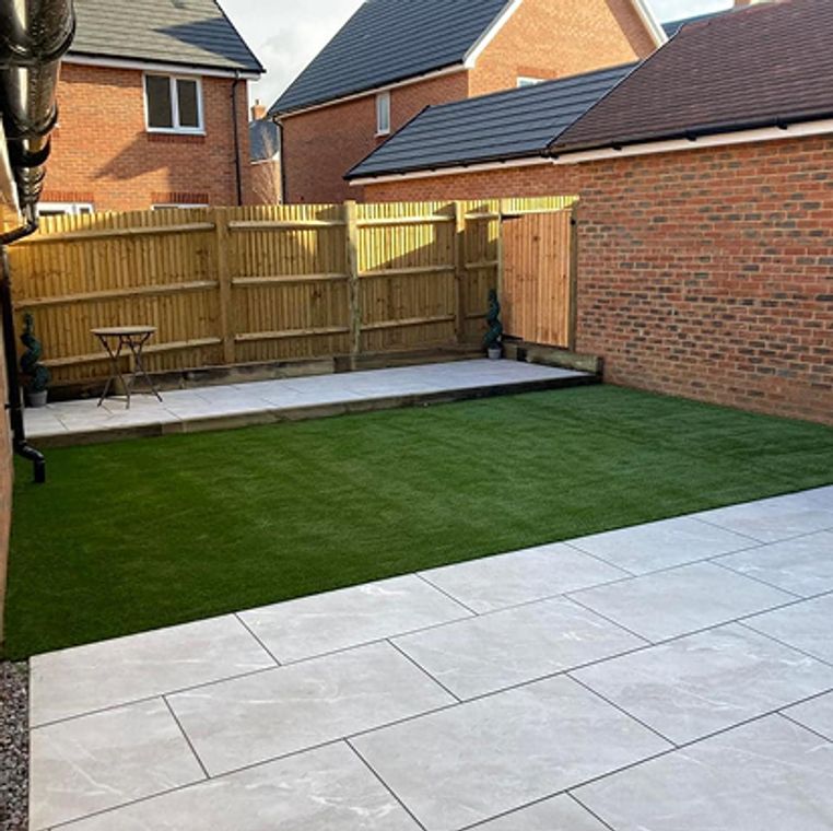 After: A landscaped garden with artificial grass, light gray patio slabs, and repaired wooden fencing.